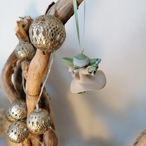 May include: A green and gray Yoda ornament hanging from a branch with silver and white decorative lights.