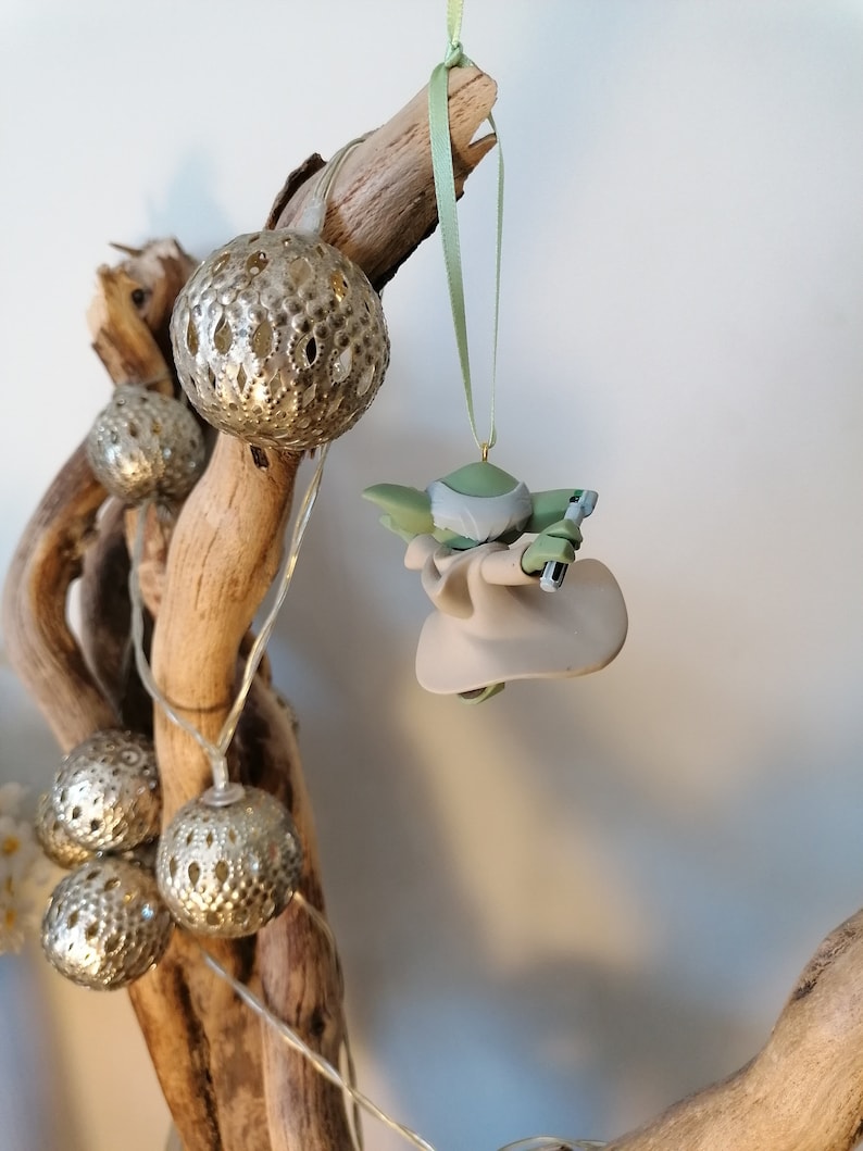May include: A green and gray Yoda ornament hanging from a branch with silver and white decorative lights.