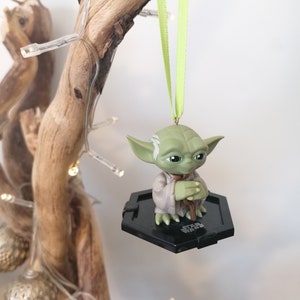 Puede incluir: Un adorno verde de Yoda colgando de una rama con una cinta verde. El adorno es una peque&ntilde;a figura de pl&aacute;stico de Yoda sentado con las manos juntas. El adorno est&aacute; sobre una base hexagonal negra con las palabras "Star Wars" impresas en ella.