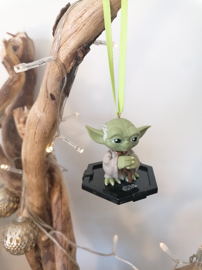 Puede incluir: Un adorno verde de Yoda colgando de una rama con una cinta verde. El adorno es una peque&ntilde;a figura de pl&aacute;stico de Yoda sentado con las manos juntas. El adorno est&aacute; sobre una base hexagonal negra con las palabras "Star Wars" impresas en ella.