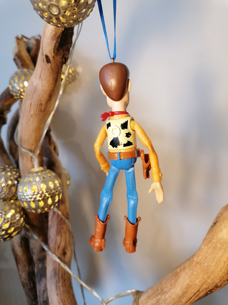 Puede incluir: Una figura de juguete de pl&aacute;stico de Woody de la franquicia de pel&iacute;culas Toy Story. La figura lleva su chaleco marr&oacute;n, sus vaqueros azules y su sombrero de vaquero caracter&iacute;sticos. Est&aacute; colgado de una cinta azul.
