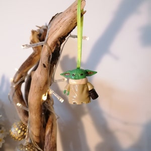 May include: A green and brown Yoda ornament hanging from a branch with a green ribbon. The ornament is a character from the Star Wars franchise.