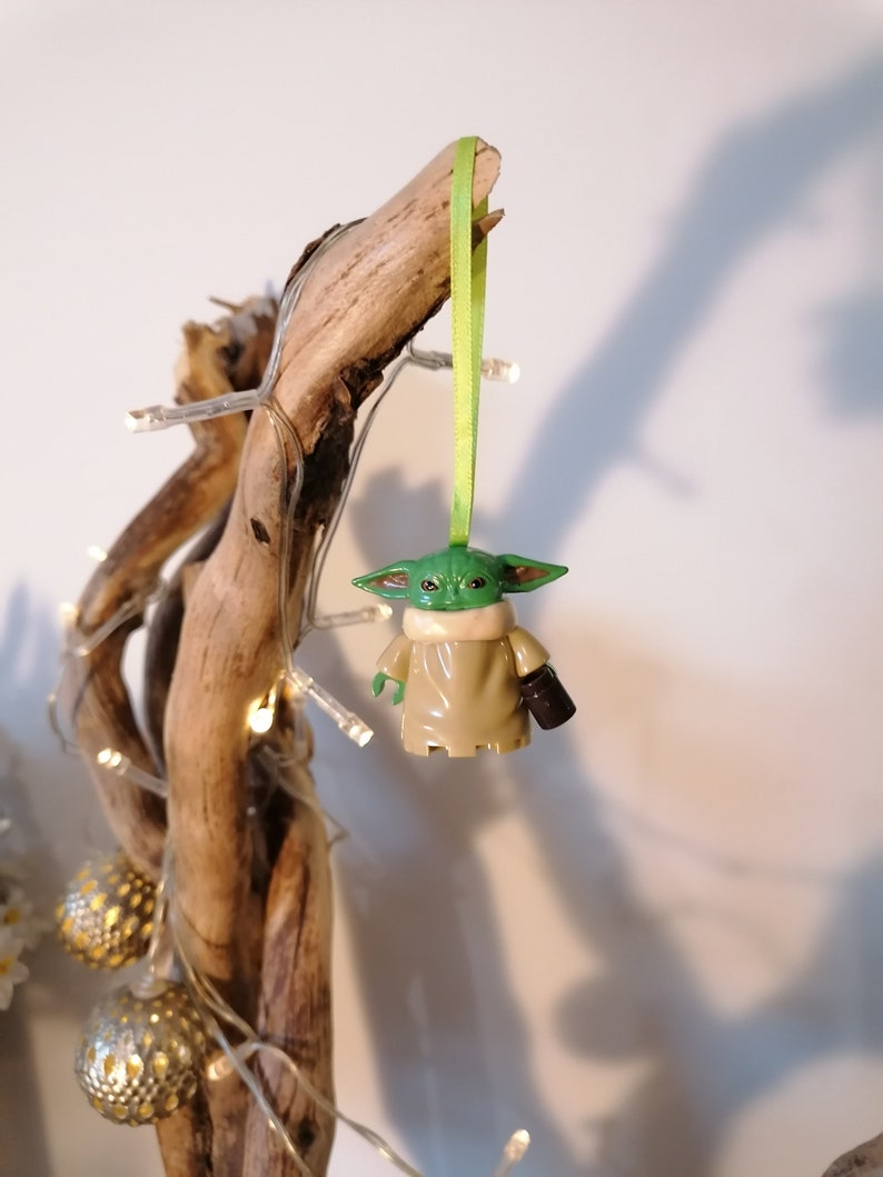 May include: A green and brown Yoda ornament hanging from a branch with a green ribbon. The ornament is a character from the Star Wars franchise.