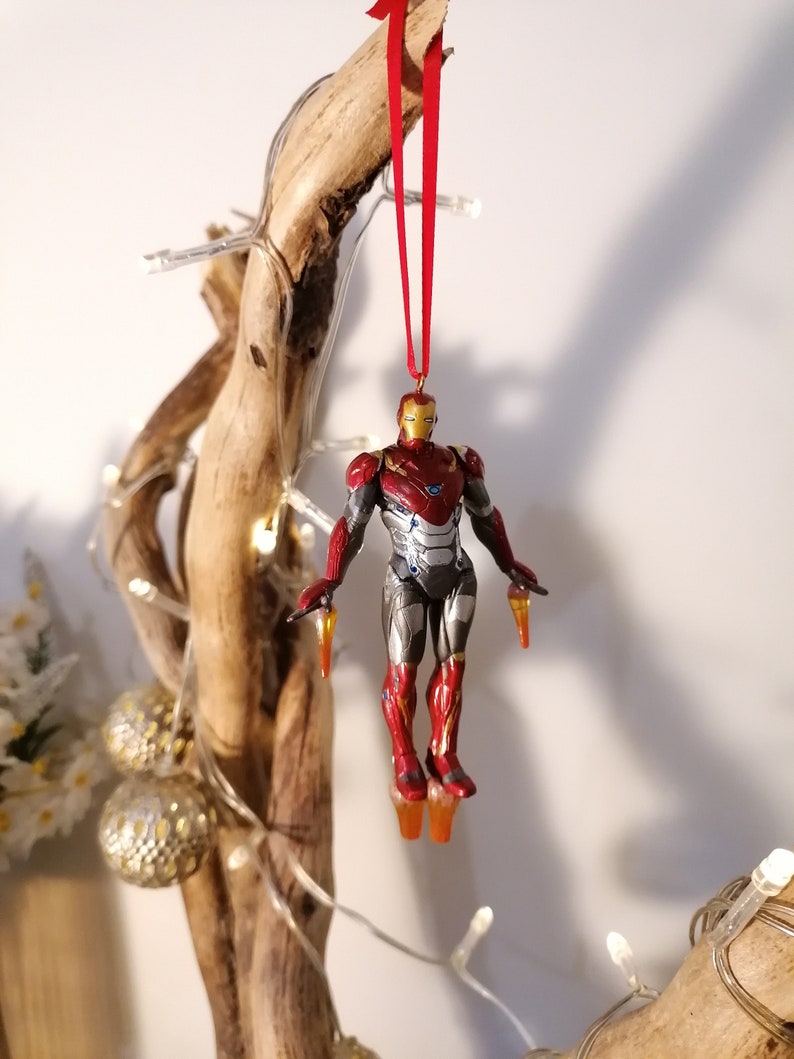 May include: A red and gold Iron Man figurine ornament hanging from a red ribbon on a wooden branch. The figurine is in a flying pose with its arms outstretched and its repulsor beams glowing.