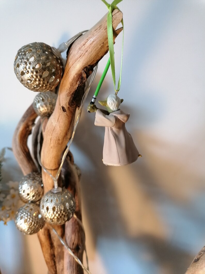 Puede incluir: Adorno navide&ntilde;o de Yoda con un sable de luz verde, suspendido de una cinta verde. La figura de Yoda es de color beige y blanco. El adorno cuelga de un trozo de madera flotante, junto a luces decorativas esf&eacute;ricas plateadas.