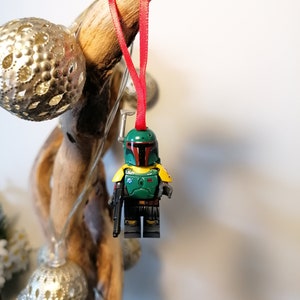 May include: A green and yellow Lego minifigure of Boba Fett, a Star Wars character, hanging from a red ribbon. The minifigure is wearing a helmet and holding a blaster.