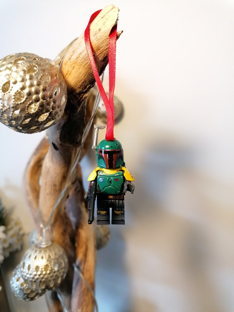 May include: A green and yellow Lego minifigure of Boba Fett, a Star Wars character, hanging from a red ribbon. The minifigure is wearing a helmet and holding a blaster.