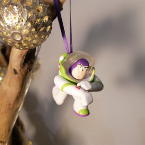 Puede incluir: Una figura de juguete de pl&aacute;stico de Buzz Lightyear de la franquicia de pel&iacute;culas Toy Story, de color morado y verde, colgando de una cinta morada. La figura est&aacute; en una pose de vuelo con un brazo levantado y el otro brazo doblado en el codo.