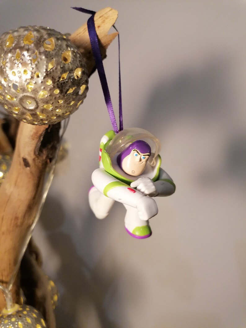 Puede incluir: Una figura de juguete de pl&aacute;stico de Buzz Lightyear de la franquicia de pel&iacute;culas Toy Story, de color morado y verde, colgando de una cinta morada. La figura est&aacute; en una pose de vuelo con un brazo levantado y el otro brazo doblado en el codo.
