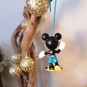 May include: A black and white Mickey Mouse ornament with blue overalls hanging from a blue ribbon. The ornament is hanging from a branch with gold string lights.