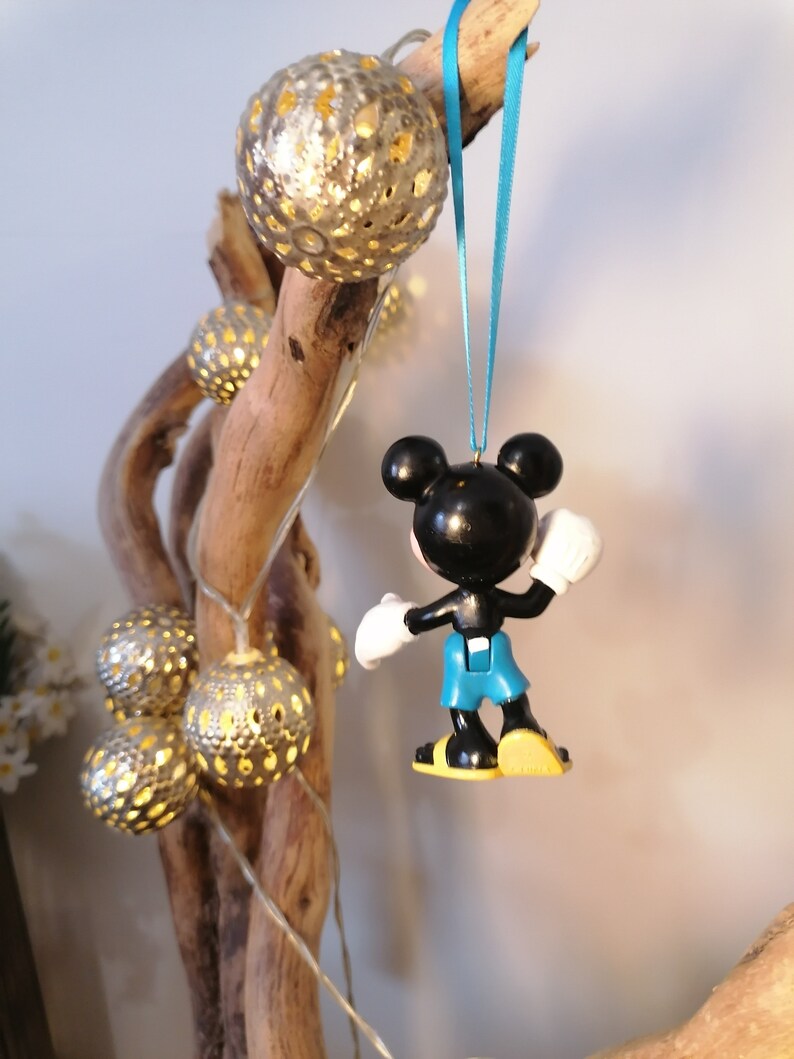 May include: A black and white Mickey Mouse ornament with blue overalls hanging from a blue ribbon. The ornament is hanging from a branch with gold string lights.