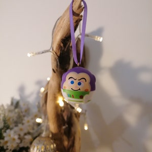 Puede incluir: Adorno navideño morado y verde con un personaje de dibujos animados con una expresión de sorpresa. El adorno está colgado de una cinta morada en una rama de madera.
