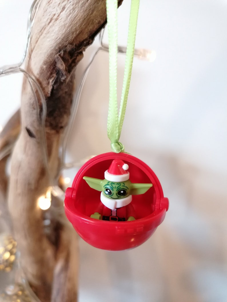 Pu&ograve; includere: Un addobbo natalizio rosso con una figura Lego verde e bianca di Baby Yoda che indossa un cappello da Babbo Natale.