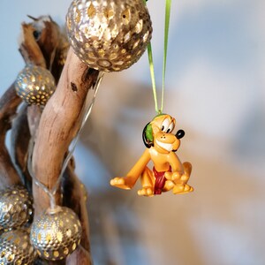 May include: A yellow cartoon dog ornament with a green leaf headband hanging from a green ribbon on a branch with gold and silver decorative balls.