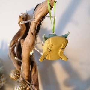 May include: A yellow and green Christmas ornament shaped like a character from the Star Wars franchise. The ornament is hanging from a green ribbon on a branch of driftwood.