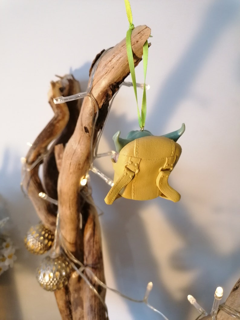 May include: A yellow and green Christmas ornament shaped like a character from the Star Wars franchise. The ornament is hanging from a green ribbon on a branch of driftwood.