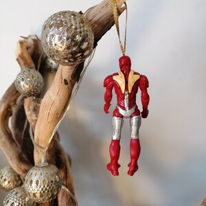 Pode incluir: Ornamento de Natal Iron Man vermelho e prateado pendurado em um galho com fita dourada e bolas prateadas.