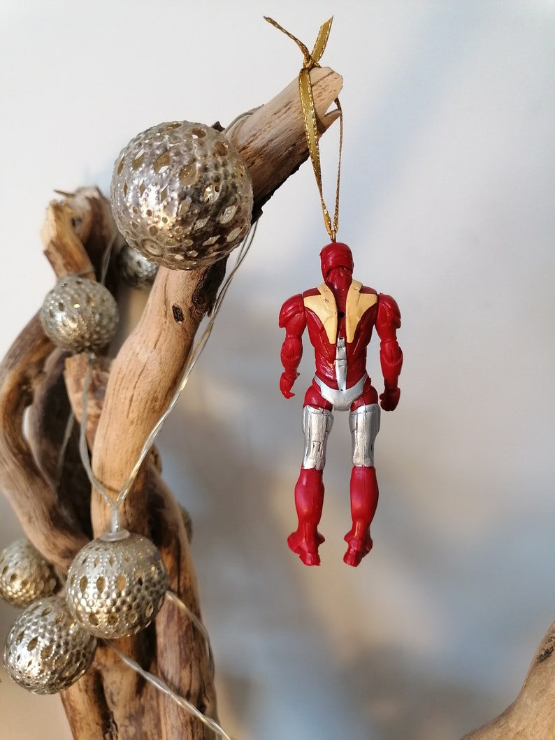 Pode incluir: Ornamento de Natal Iron Man vermelho e prateado pendurado em um galho com fita dourada e bolas prateadas.