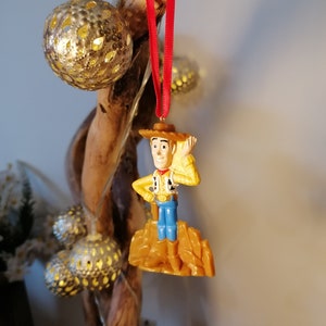 May include: A red string ornament featuring a plastic figurine of Woody from Toy Story. Woody is wearing his signature cowboy hat, vest, and jeans. He is standing on a brown plastic rock.