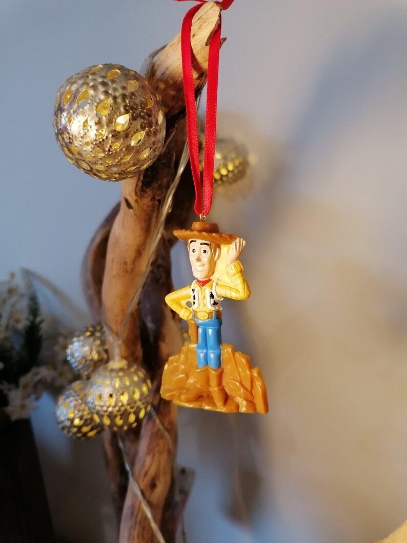 May include: A red string ornament featuring a plastic figurine of Woody from Toy Story. Woody is wearing his signature cowboy hat, vest, and jeans. He is standing on a brown plastic rock.