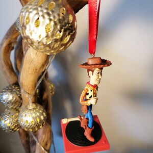 Puede incluir: Un adorno navide&ntilde;o rojo y negro con una figura de Woody de Toy Story de pie sobre un tocadiscos. Woody lleva su caracter&iacute;stico sombrero de vaquero, chaleco y botas.
