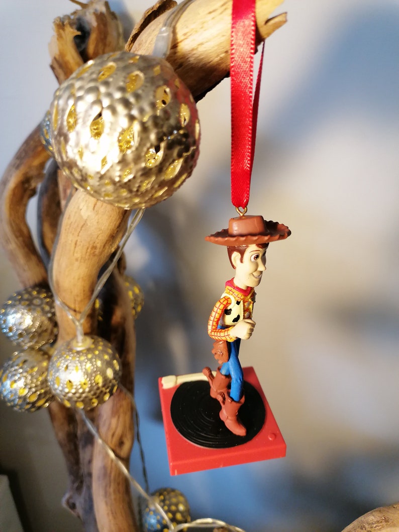 Puede incluir: Un adorno navide&ntilde;o rojo y negro con una figura de Woody de Toy Story de pie sobre un tocadiscos. Woody lleva su caracter&iacute;stico sombrero de vaquero, chaleco y botas.