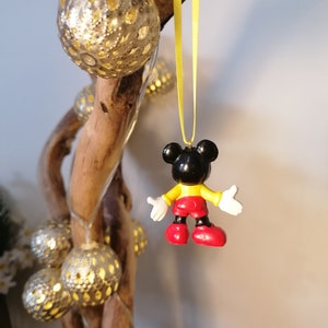 K&ouml;nnte beinhalten: Ein schwarzer und gelber Mickey Mouse-Anh&auml;nger, der an einem gelben Band h&auml;ngt. Der Anh&auml;nger ist aus Kunststoff und hat ein rot-gelbes Outfit. Der Anh&auml;nger h&auml;ngt an einem Ast mit silbernen und wei&szlig;en Dekorationslichtern.