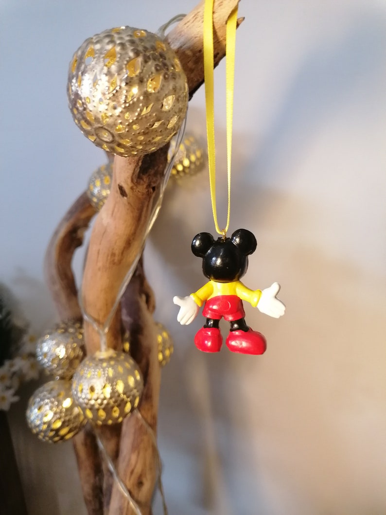 K&ouml;nnte beinhalten: Ein schwarzer und gelber Mickey Mouse-Anh&auml;nger, der an einem gelben Band h&auml;ngt. Der Anh&auml;nger ist aus Kunststoff und hat ein rot-gelbes Outfit. Der Anh&auml;nger h&auml;ngt an einem Ast mit silbernen und wei&szlig;en Dekorationslichtern.