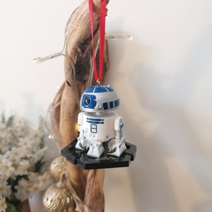 May include: A white and blue R2-D2 Star Wars character ornament hanging from a red ribbon on a piece of driftwood. The ornament is on a black base.