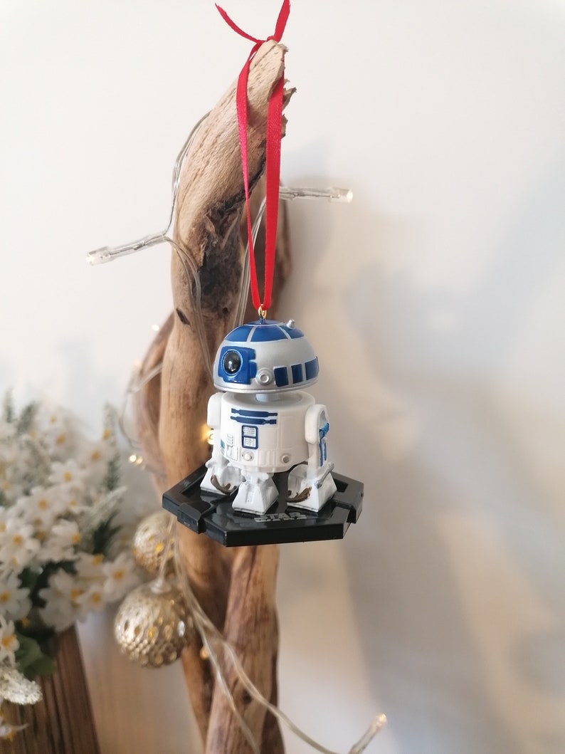 May include: A white and blue R2-D2 Star Wars character ornament hanging from a red ribbon on a piece of driftwood. The ornament is on a black base.