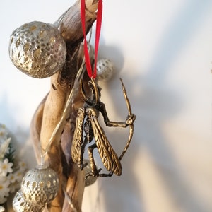 May include: A gold-coloured ornament of a winged creature holding a bow and arrow, hanging from a red ribbon on a branch.