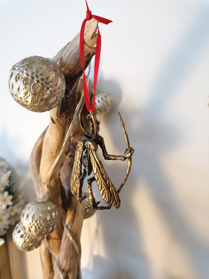 May include: A gold-coloured ornament of a winged creature holding a bow and arrow, hanging from a red ribbon on a branch.