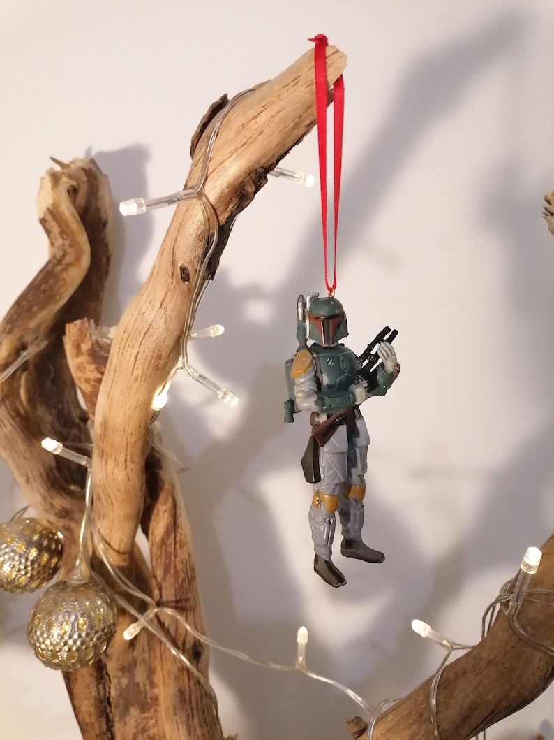 May include: A green and gray Star Wars Boba Fett action figure ornament hanging from a branch with a red ribbon. The ornament is decorated with a brown belt and yellow accents.