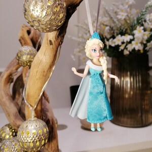 May include: A blue and white plastic doll of Elsa from the movie Frozen hanging from a branch with a string. The doll is wearing a blue dress with a white cape and has blonde hair. The branch is decorated with gold metallic ornaments.