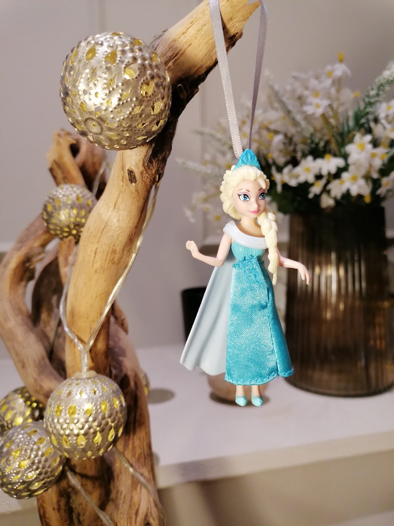 May include: A blue and white plastic doll of Elsa from the movie Frozen hanging from a branch with a string. The doll is wearing a blue dress with a white cape and has blonde hair. The branch is decorated with gold metallic ornaments.