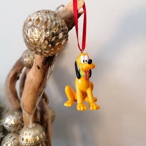 Op de afbeelding: Een gele en bruine cartoonhond kerstversiering die aan een rood lint hangt. De versiering is bevestigd aan een tak met gouden en witte decoratieve ballen.