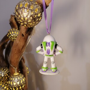 Puede incluir: Un adorno navide&ntilde;o de Buzz Lightyear blanco y verde con una pieza de pecho morada y una base blanca. El adorno est&aacute; colgado de una cinta morada.