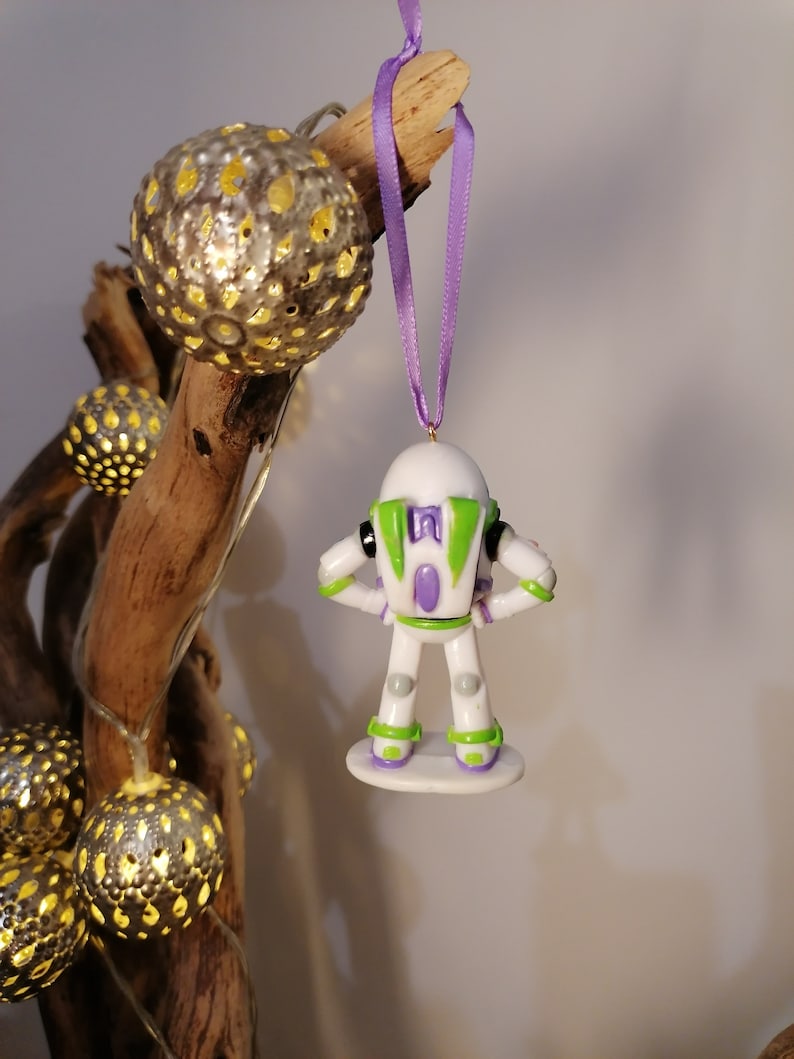 Puede incluir: Un adorno navide&ntilde;o de Buzz Lightyear blanco y verde con una pieza de pecho morada y una base blanca. El adorno est&aacute; colgado de una cinta morada.
