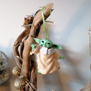 May include: A green and beige Christmas ornament featuring a character from the Star Wars franchise, The Mandalorian. The ornament is hanging from a branch with a green ribbon.