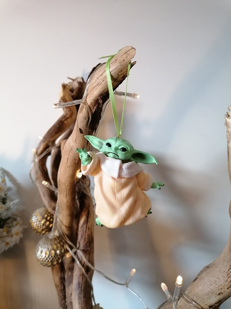 May include: A green and beige Christmas ornament featuring a character from the Star Wars franchise, The Mandalorian. The ornament is hanging from a branch with a green ribbon.