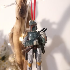 May include: A green and gray Star Wars Boba Fett Christmas ornament with a red string hanging from the top. The ornament is a plastic figurine of Boba Fett holding his blaster rifle.