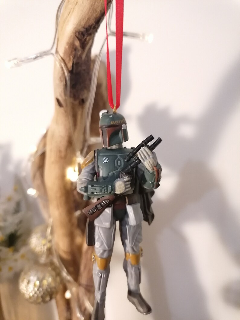May include: A green and gray Star Wars Boba Fett Christmas ornament with a red string hanging from the top. The ornament is a plastic figurine of Boba Fett holding his blaster rifle.