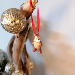 Pode incluir: Uma pequena figura de madeira marrom e bege de um esquilo de desenho animado pendurado em uma fita vermelha. O esquilo est&aacute; usando uma gravata borboleta vermelha e tem a barriga branca. A figura est&aacute; pendurada em um galho de uma &aacute;rvore.