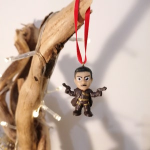 May include: A brown and black figurine of a man holding two guns, hanging from a red ribbon on a branch. The figurine is a Christmas ornament.