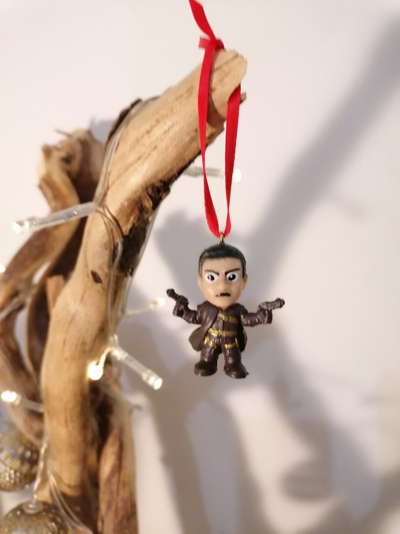May include: A brown and black figurine of a man holding two guns, hanging from a red ribbon on a branch. The figurine is a Christmas ornament.