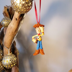 Pu&ograve; includere: Uno spago rosso pende da un ramo con una statuetta di plastica di Woody di Toy Story. Woody indossa il suo cappello da cowboy, il gilet e i jeans caratteristici. Si sta tenendo la mano destra sulla testa e sorride.