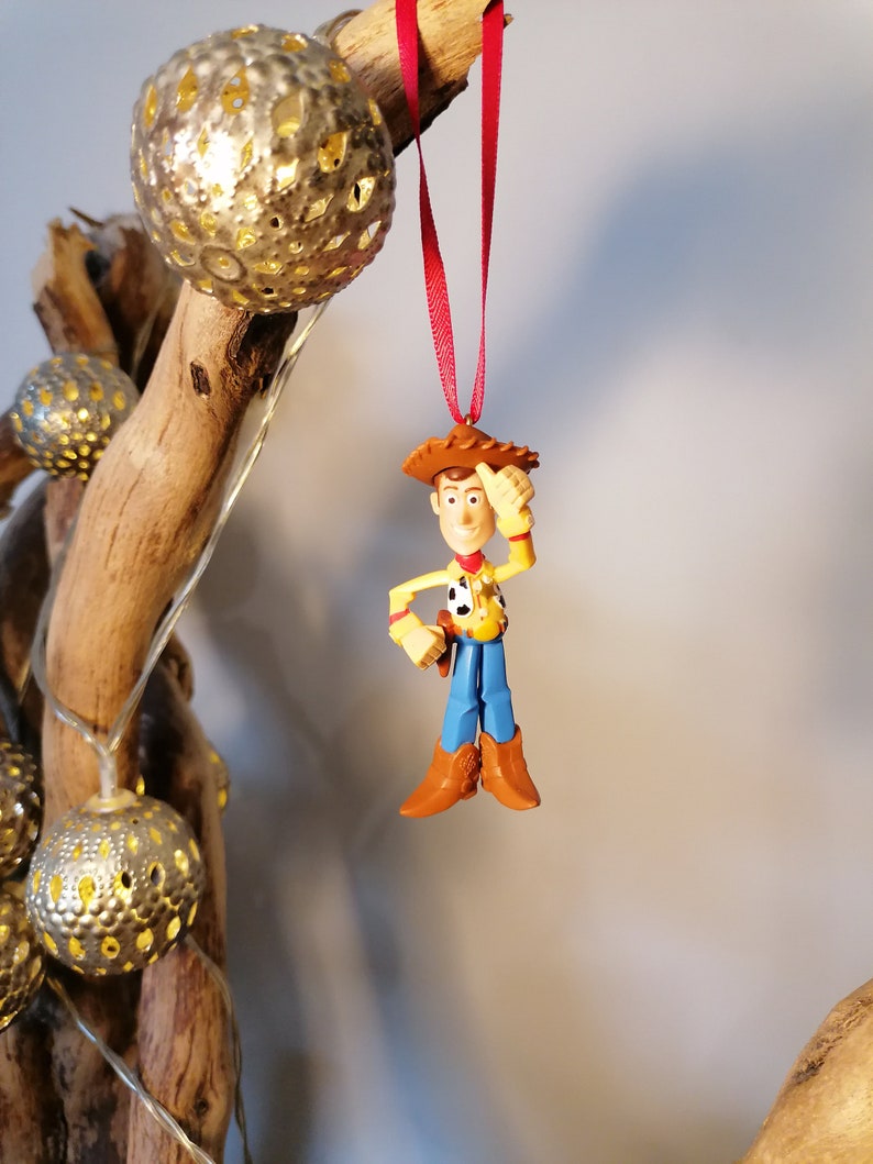 Pu&ograve; includere: Uno spago rosso pende da un ramo con una statuetta di plastica di Woody di Toy Story. Woody indossa il suo cappello da cowboy, il gilet e i jeans caratteristici. Si sta tenendo la mano destra sulla testa e sorride.
