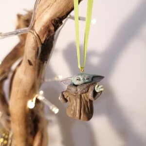 Pu&ograve; includere: Una piccola statuina verde e marrone di un personaggio del franchise di Star Wars, appesa a un nastro verde su un ramo. La statuina indossa un cappotto marrone e ha grandi orecchie.