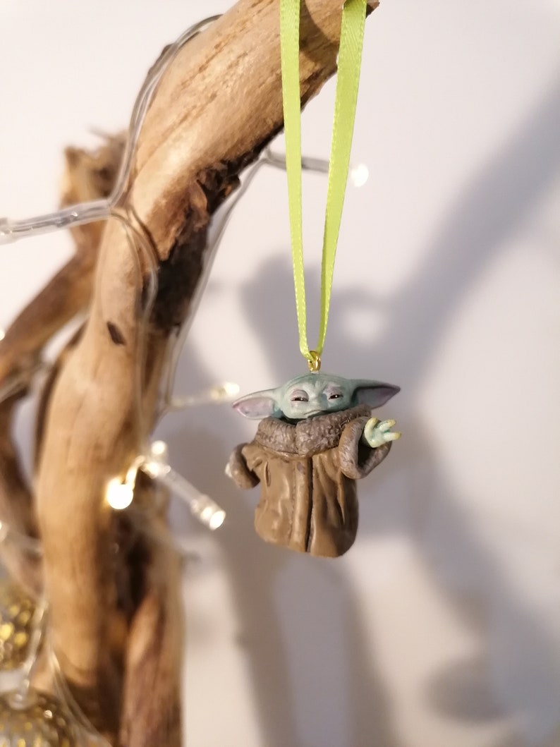 Pu&ograve; includere: Una piccola statuina verde e marrone di un personaggio del franchise di Star Wars, appesa a un nastro verde su un ramo. La statuina indossa un cappotto marrone e ha grandi orecchie.
