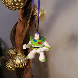 May include: A white, green and purple plastic ornament of Buzz Lightyear from the Toy Story franchise. The ornament is hanging from a purple ribbon.